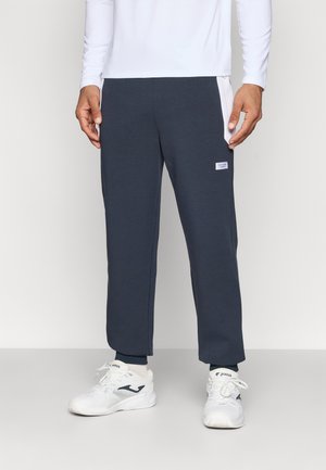 Tumesin sinine sportlik jooksupükste mudel, millel on kitsenev lõige, elastne vöökoht ja külgmised võrkpaneelid. Kombineeritud valgete tossudega.