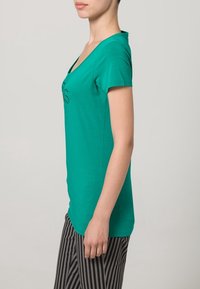 Camiseta verde de manga corta con escote en V y un patrón decorativo, combinada con pantalones de rayas en blanco y negro. Textura de algodón suave.