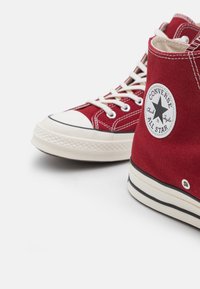 Sapatilhas altas de lona vermelha com solas de borracha brancas, detalhe de costura clássica e um emblema circular com uma estrela preta.