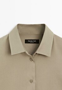 Overhemd met knopen van lichtbeige stof, met een klassieke kraag, sluiting met zes knopen en een merkembleem aan de binnenkant van de kraag.