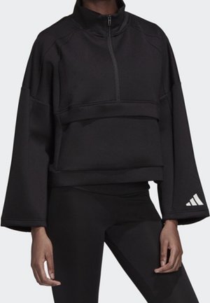 Czarna bluza z długim rękawem, zapinana na pół suwaka, o szerokich rękawach, noszona z czarnymi legginsami, z białym logo Adidas na lewym mankiecie.