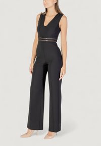 Mono negro con escote en V profundo, detalle dorado en la cintura y diseño de pierna ancha. Tela suave y ajuste entallado. Tacones nude a juego.