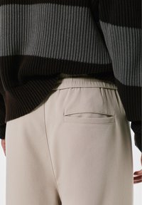 Smeđa i siva prugasta rebrasta pletiva kombinirana s laganim bež hlačama koje imaju jedan stražnji džep i elastičnu traku u struku.