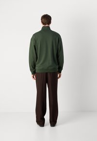 Felpa verde con collo alto, polsini e orlo a coste. Abbinata a pantaloni marroni larghi. Design semplice e funzionale.
