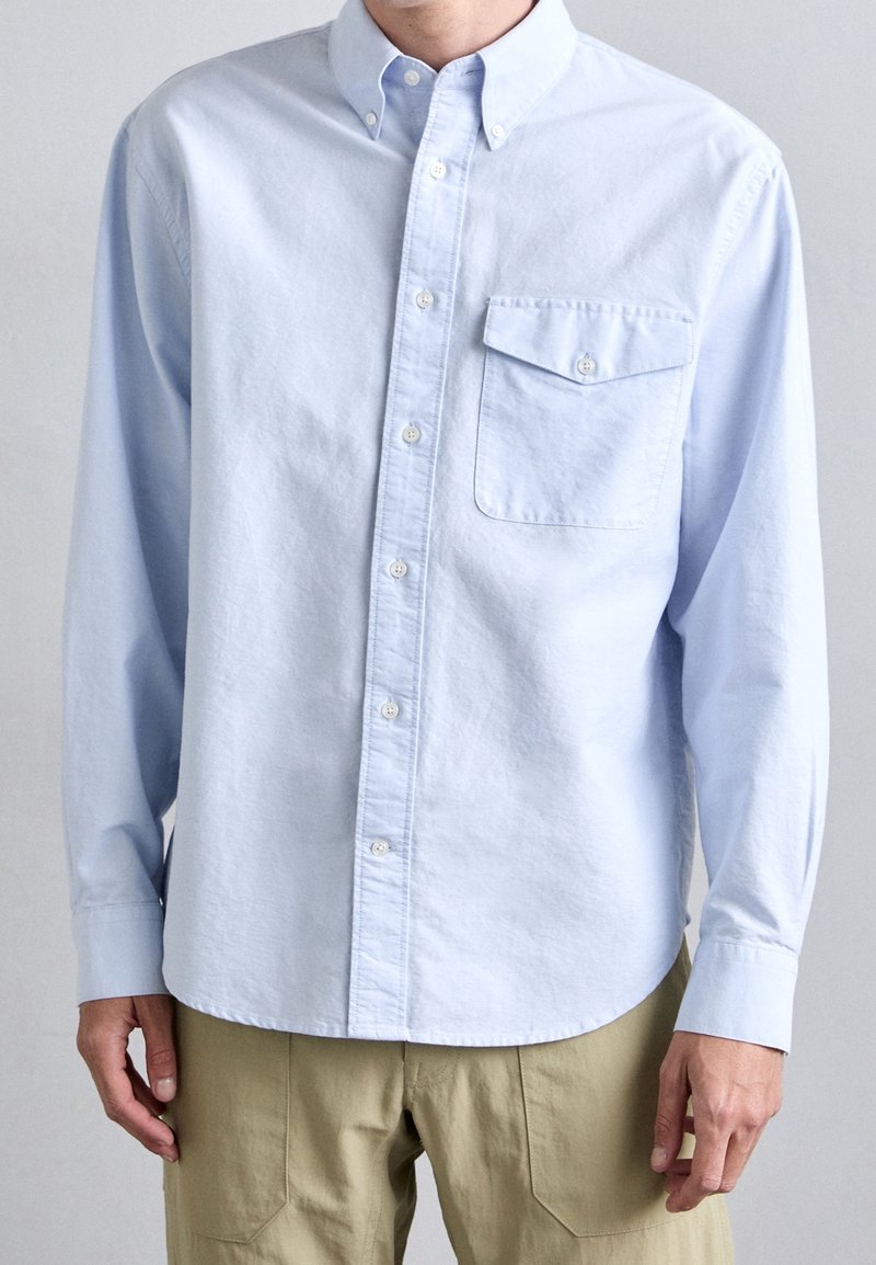 Homme portant une chemise bleu clair boutonnée avec une grande poche avant et un pantalon beige, debout devant un fond uni.