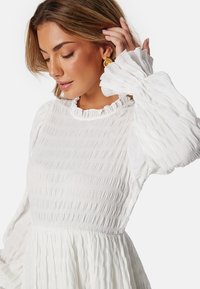Biała, teksturowana sukienka z marszczonym korpusem, falbaniastym dekoltem i bufiastymi, plisowanymi rękawami. Charakteryzuje się lekkim, zwiewnym fasonem.