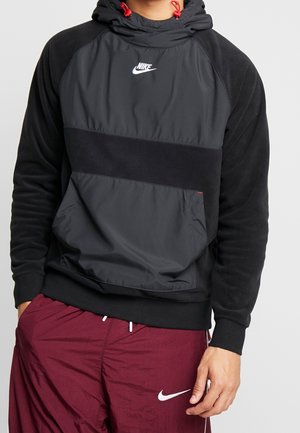 Person trägt einen schwarzen und dunkelgrauen Nike-Hoodie mit roten Kordeln und bordeauxrote Nike-Sportshorts mit weißem Logo.