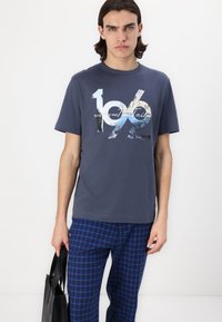 Mladić u tamnoplavoj majici s grafikom i tekstom "1962" te plavim kariranim hlačama, drži crnu torbu na bijeloj pozadini.