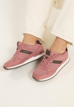 Person iført lyserøde mesh- og ruskindssneakers med brune accenter og hvide såler, kombineret med off-white bukser.