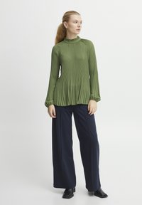 Blusa verde a pieghe con collo alto e maniche lunghe, abbinata a pantaloni larghi blu navy e stivali neri con tacco alla caviglia. Texture liscia.
