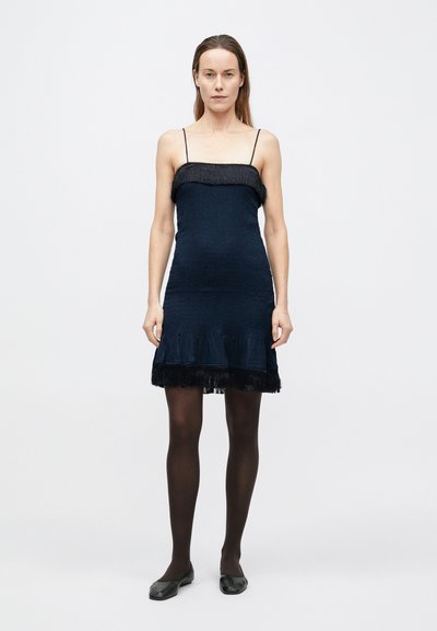 Tengerészkék ruha spagettipántokkal, texturált anyagból, rojtos szegéllyel, testhezálló és bővülő aljrészel. Fekete harisnya és lapos cipő.