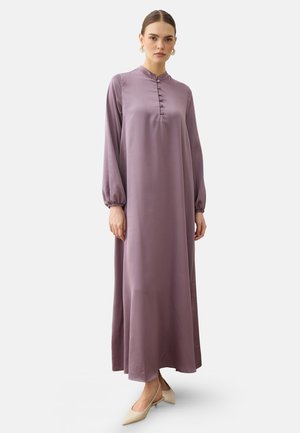 Frau, die ein langes mauvefarbenes, geknöpftes Kleid mit langen Ärmeln und beigefarbenen, flachen spitzen Schuhen trägt, steht vor einem einfarbigen Hintergrund.