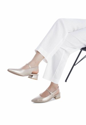 Zapatos slingback plateados metálicos con punta puntiaguda, tacón grueso y correa en el tobillo, combinados con pantalones blancos de pierna ancha.
