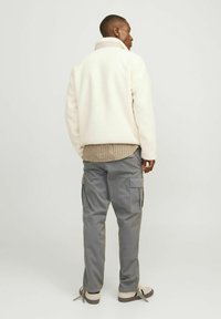 Fleece bunda v off-white farbe s vysokým golierom, preložená cez hnedú kockovanú košeľu; skombinovaná so sivými cargo nohavicami so bočnými vreckami.