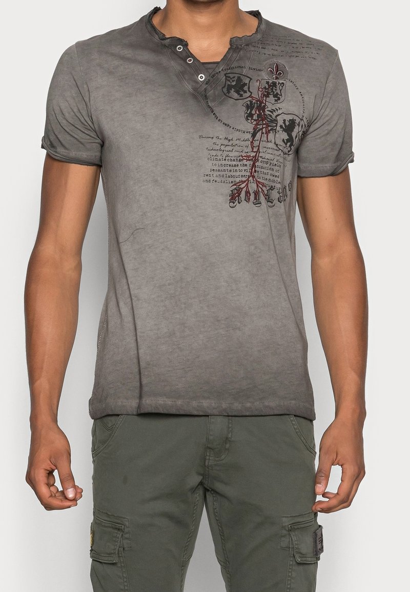 Camiseta gris de manga corta con un cuello tipo henley, que presenta una textura desvanecida, gráficos impresos y detalles de dobladillo desgastado.