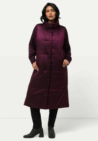 Langer, pflaumenfarbener Puffer-Westen mit hohem Kragen, Rautensteppung und seitlichen Taschen, getragen über einem schwarzen Langarmtop und einer Hose.