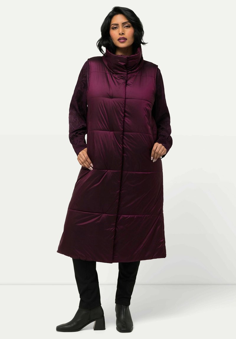 Langer, pflaumenfarbener Puffer-Westen mit hohem Kragen, Rautensteppung und seitlichen Taschen, getragen über einem schwarzen Langarmtop und einer Hose.
