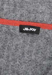 Grijze stof met een gestructureerd, fuzzy oppervlak, voorzien van een helderrode accentstreep en een zwarte label met de tekst "J&Joy."
