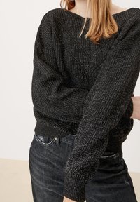 Czarny sweter wykonany na drutach z luźnym krojem, dekoltem w łódź, strukturalnym wzorem i ściągaczami na rękawach. Noszony z ciemnymi dżinsami.