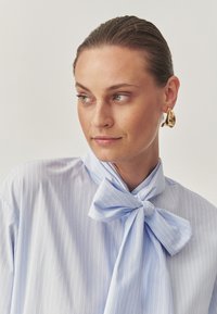 Light blue pinstriped blouse with a bow tie collar, made of smooth fabric. Features a relaxed fit and subtle sheen. Gold statement earring accent.