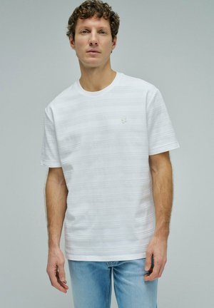 Hombre con cabello rizado que lleva una camiseta blanca lisa con textura y mangas cortas y vaqueros azul claro, de pie sobre un fondo gris.