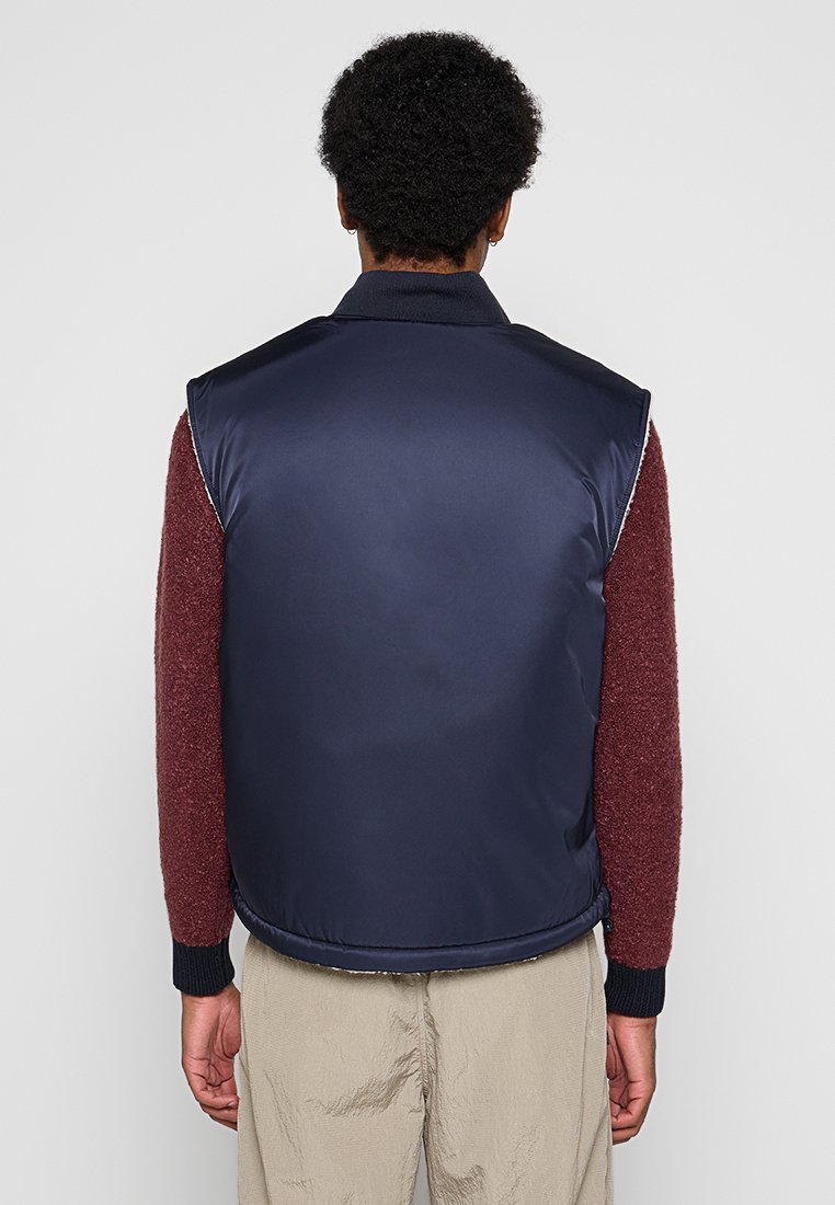 Gilet imbottito blu navy con colletto alto, design senza maniche e texture trapuntata, indossato sopra un maglione bordeaux.