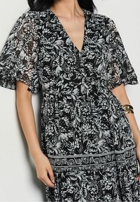 Svart blommig klänning med V-ringning, korta volangärmar och intrikata vita bladmönster. Lätt, flödande tyg med elastisk midja.