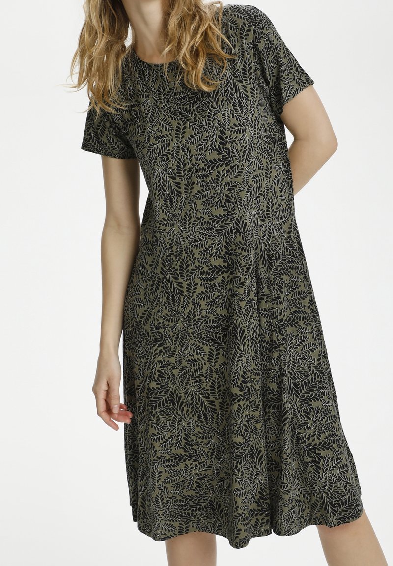 Femme portant une robe verte à manches courtes, longueur genou, avec un motif feuille noire sur fond blanc.
