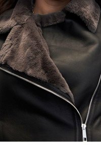Zbliżenie na czarną skórzaną kurtkę z szerokim brązowym sztucznym futrem na kołnierzu i srebrnym zamkiem błyskawicznym częściowo rozpiętym z przodu.