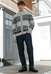 Strikket cardigan i grå med sorte geometriske mønstre, lynlås lukning, parret med mørkeblå jeans og mørke støvler på en stenoverflade.