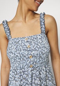 Femme souriante portant une robe bleue sans manches avec un motif floral blanc et bleu clair et des boutons en bois à l'avant.