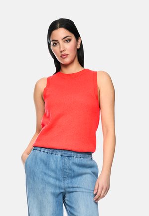 Frau mit glatten schwarzen Haaren, die ein ärmelloses, hellrotes Stricktop und hellblaue Jeans trägt, steht mit den Händen in den Taschen.