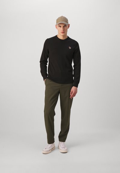 Μαύρο φούτερ με στρογγυλή λαιμόκοψη και μικρό κεντημένο λογότυπο, συνδυασμένο με ελαιοπράσινα παντελόνια και ανοιχτόχρωμα αθλητικά παπούτσια. Μπέζ καπέλο στο κεφάλι.