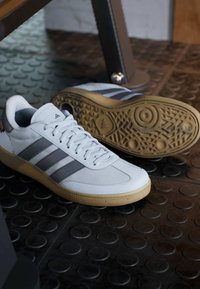 Witte sportieve schoenen met grijze strepen, een gestructureerde mesh bovenzijde, rubberen zool en een ronde neus. Zichtbaar profielpatroon op de zool.