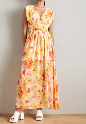 Robe maxi sans manches à imprimé floral jaune, orange et rose, décolleté en V profond, taille cintrée, associée à des mules blanches à talons.