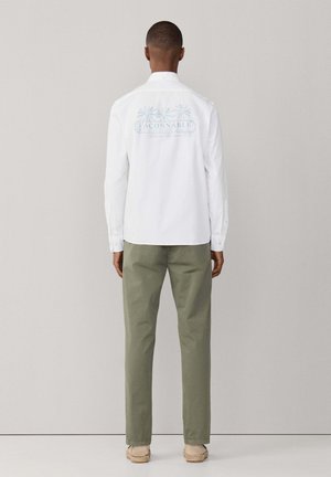 Homme debout de dos portant une chemise à manches longues blanche avec un motif de palmiers bleus et un pantalon vert, sur un fond uni.