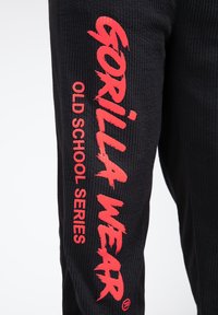 Sorte bukser med en tekstureret overflade, prydet med fed rød tekst "GORILLA WEAR" og "OLD SCHOOL SERIES" på siden.