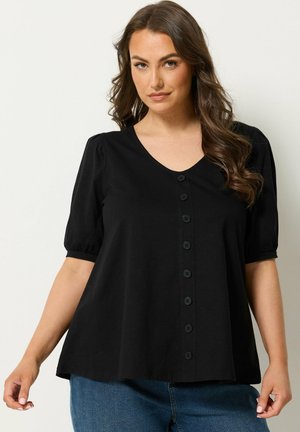 Femme aux longs cheveux ondulés portant un haut noir à manches courtes boutonné et un jean bleu, debout devant un fond clair uni.