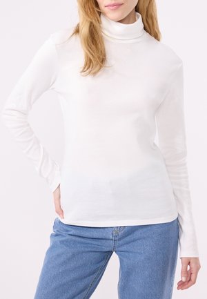 Frau in einem weißen langärmeligen Rollkragenoberteil und blauen Jeans, mit einer Hand auf ihrer Hüfte vor einem schlichten hellen Hintergrund.