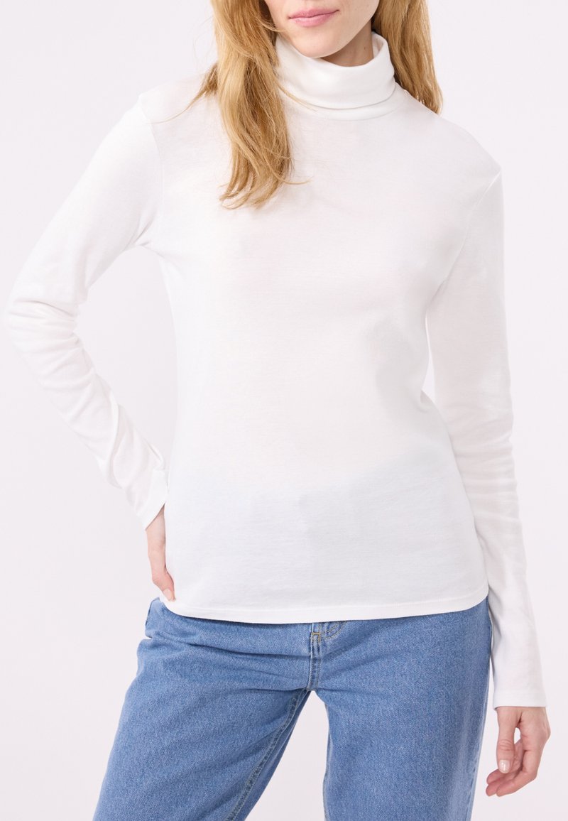 Vrouw in een witte lange mouwen coltrui en blauwe spijkerbroek, met één hand op haar heup tegen een effen lichte achtergrond.