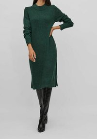 Abito verde a maglia con scollatura rotonda, maniche lunghe, spacchi laterali e vestibilità comoda, abbinato a stivali neri lucidi al ginocchio.