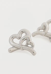 Σκουλαρίκια σε σχήμα καρδιάς από μέταλλο σε ασημί απόχρωση με διαπλεκόμενες σχεδιαστικές λεπτομέρειες. Διαθέτουν εγχάρακτη επωνυμία "GUESS", λεία υφή και γυαλιστερό φινίρισμα.