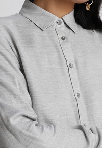 Grå skjorta med knappar gjord av texturerad tyg, med spetsig krage och knappstängning. Långa ärmar synliga, med subtilt fiskbensmönster.