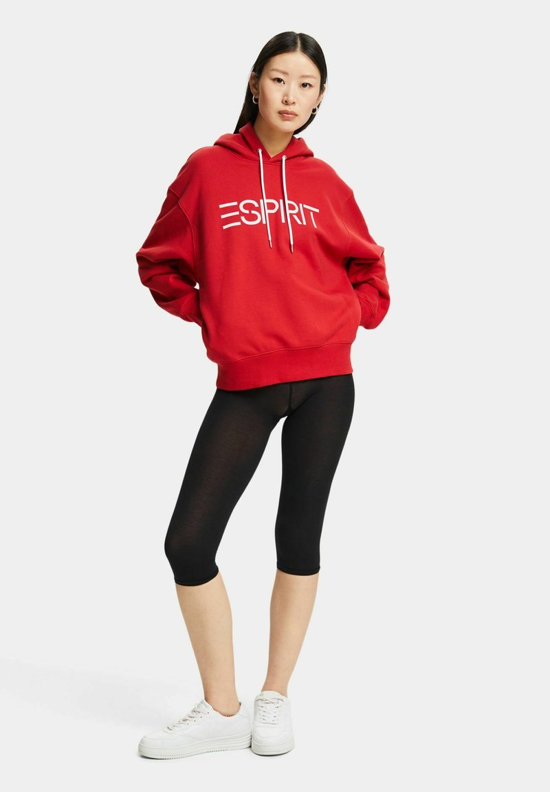 Rode hoodie met het witte "ESPRIT" logo, ruime pasvorm, geribde manchetten, gecombineerd met zwarte leggings en witte sneakers. Effen achtergrond.