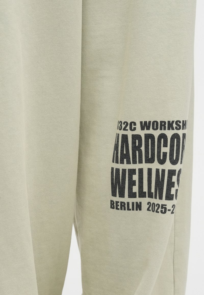 Lichtgroene sweatshirt van katoen met gedurfde zwarte bedrukking. De tekst omvat "032C WORKSHOP HARDCORE WELLNESS BERLIJN 2025-2026."