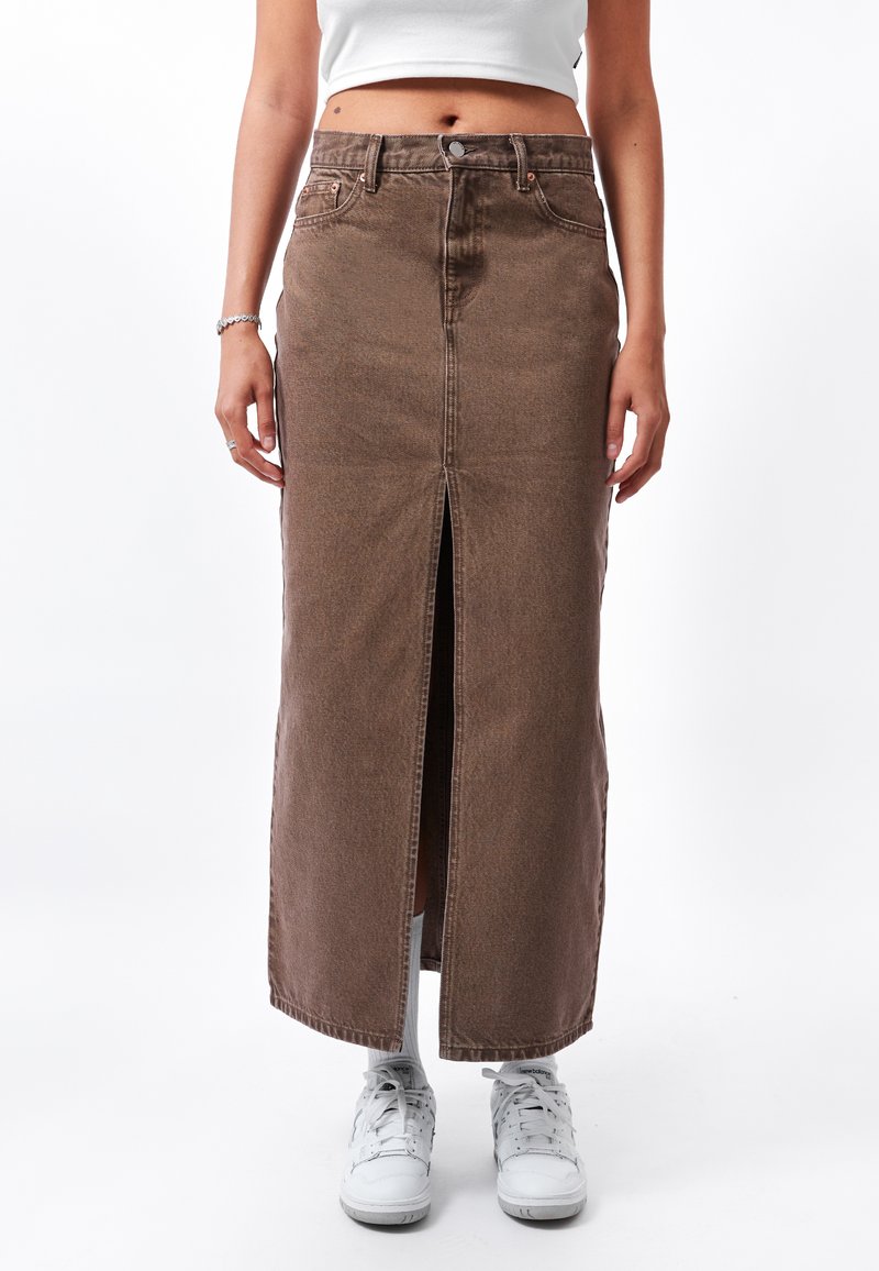 Falda maxi de mezclilla marrón con una abertura frontal, cintura alta y costuras en contraste. Combinada con zapatillas blancas y calcetines tobilleros.