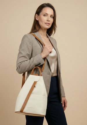 Kvinde iført beige blazer og mørke bukser holder en hvid og brun læderhåndtaske med skulderstrop mod en beige baggrund.