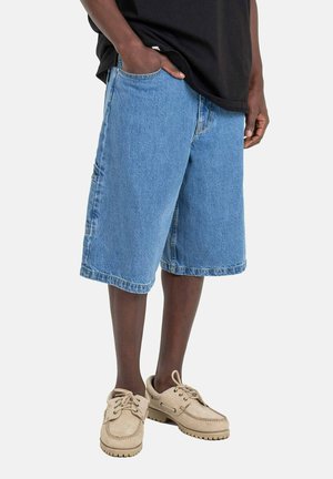 Jeansshorts aus leicht blauer Stoff, mit lockerer Passform, klassischen Taschen und umgeschlagenem Saum. Getragen mit beigen Schuhen und einem schwarzen Shirt.