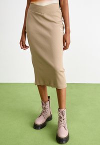 Falda midi de canalé beige con diseño de torsión en la parte frontal y detalle del logo, combinada con botas rosas claro con cordones, suelas negras y acentos amarillos.