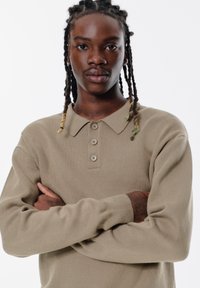 Khaki polo tričko s límečkem, zapínáním na tři knoflíky a dlouhými rukávy. Texturovaná tkanina s uvolněným střihem.